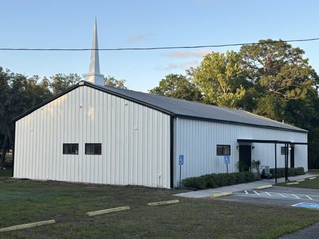 The front of the church on a sunny day: white metal siding, black trim, a tall white steeple