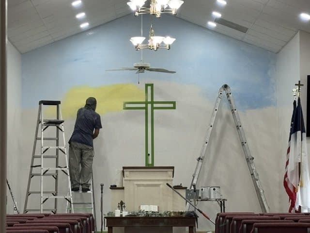 A member on a ladder painting the sky of the mural behind the pulpit