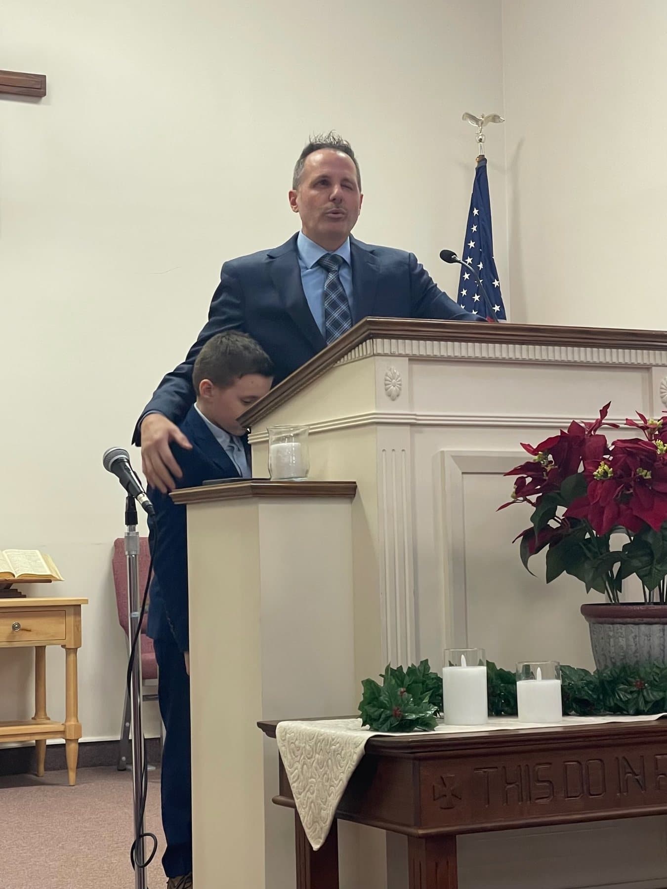 Pastor Bankert at the pulpit with a child beside him, poinsettias on the platform
