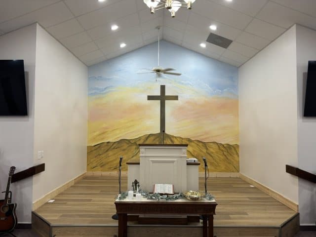 The hand-painted mural behind the pulpit: a cross on a golden hill under a sunrise sky